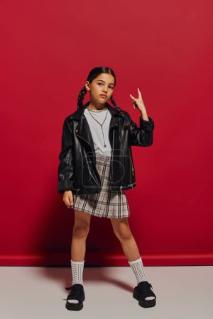 Full length of fashionable preteen girl with hairstyle wearing leather jacket and plaid skirt while showing rock sign at camera and standing on red background, stylish preteen outfit concept