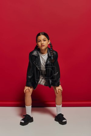 Full length of fashionable and confident preteen girl in leather jacket and plaid skirt looking at camera while posing and standing on red background, stylish preteen outfit concept