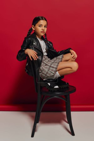 Fashionable preadolescent girl with hairstyle wearing leather jacket and plaid skirt while looking at camera and posing on chair on red background, stylish preteen outfit concept