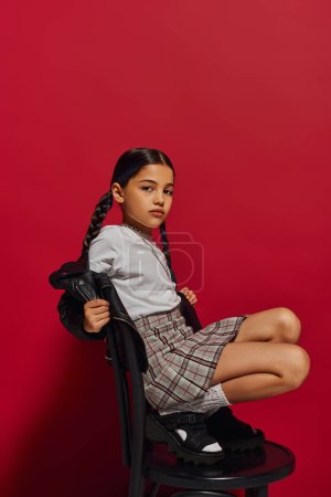 Trendy preteen Mädchen mit Frisur posiert in Lederjacke und kariertem Rock und schaut in die Kamera, während sie auf einem Stuhl auf rotem Hintergrund sitzt, stilvolles preteen Outfit-Konzept