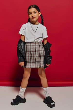 Photo for Full length of trendy brunette preteen girl with hairstyle posing in plaid skirt and leather jacket while looking at camera and standing on red background, stylish preteen outfit concept - Royalty Free Image