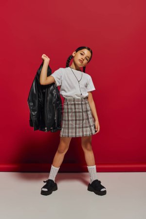 Brunette preadolescent girl with hairstyle holding leather jacket while posing in checkered skirt and looking at camera while posing on red background, stylish preteen outfit concept