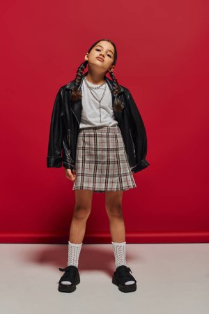 Full length of confident and fashionable preteen girl with hairstyle wearing leather jacket and plaid skirt while standing and posing on red background, stylish preteen outfit concept