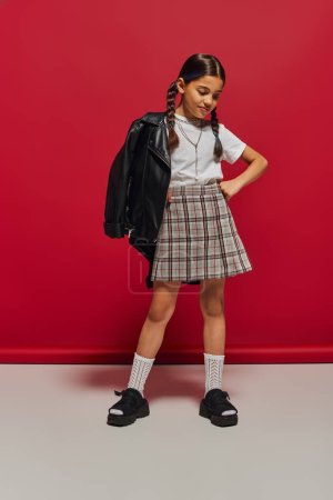Trendiges brünettes Preteen-Mädchen mit Frisur posiert in Lederjacke und kariertem Rock, hält die Hand an der Hüfte und steht auf rotem Hintergrund, stilvolles Preteen-Outfit-Konzept