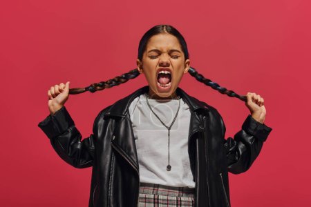 Mad and fashionable preadolescent girl screaming and touching hairstyle while posing in leather jacket and white t-shirt and standing isolated on red, girl with cool and contemporary look