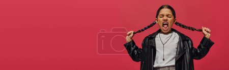Mad and screaming preteen girl in fashionable leather jacket touching hairstyle while posing and standing isolated on red, girl with cool and contemporary look, banner with copy space