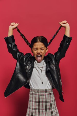 Mad preadolescent girl sticking out tongue and holding hairstyle while posing in leather jacket and plaid skirt and standing on red background, girl with cool and contemporary look