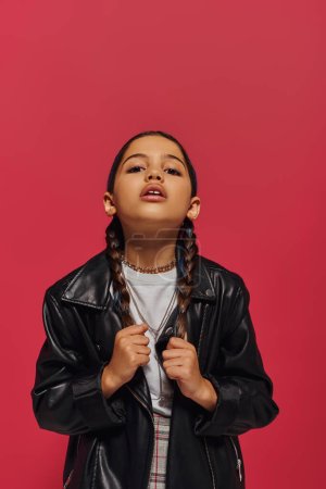 Porträt einer modischen Frühchen-Mädchen, die Haare berührt, während sie in moderner Lederjacke posiert und in die Kamera schaut und isoliert auf rot steht, Mädchen mit coolem und zeitgenössischem Look