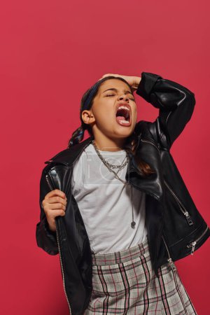 Angry and fashionable preteen girl with hairstyle screaming and touching head while posing in leather jacket and standing isolated on red, hairstyle and trendy accessories concept
