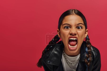 Portrait of mad preadolescent girl with hairstyle wearing leather jacket while screaming at camera and standing and posing isolated on red, hairstyle and trendy accessories concept
