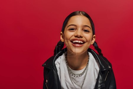 Photo for Portrait of pleased preadolescent girl with hairstyle wearing stylish leather jacket and looking at camera while posing isolated on red, hairstyle and trendy accessories concept - Royalty Free Image