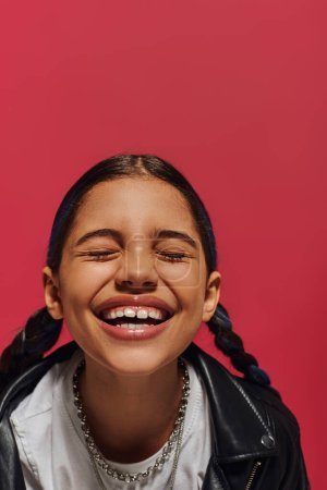 Porträt eines lächelnden Frühchens mit Frisur, die Halsketten und Lederjacke trägt, während sie mit geschlossenen Augen steht und isoliert auf Rot posiert, Frisur und trendiges Accessoire-Konzept