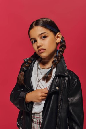 Portrait of confident and trendy preadolescent girl with dyed hair and hairstyle posing in necklaces and leather jacket while standing isolated on red, hairstyle and trendy accessories concept