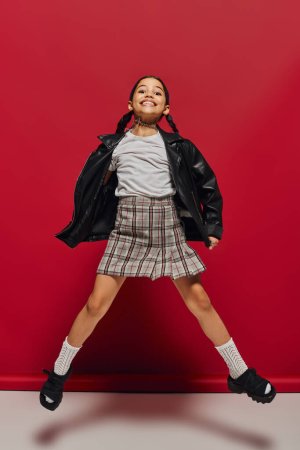 Full length of cheerful preteen kid in stylish leather jacket and plaid skirt looking at camera while jumping and having fun on red background, hairstyle and trendy accessories concept