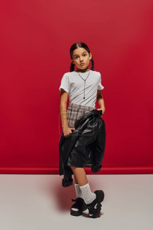 Preadolescent and fashionable girl with hairstyle wearing plaid skirt and holding leather jacket while looking at camera and posing on red background, hairstyle and trendy accessories concept