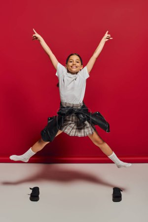 Cheerful and trendy preteen girl in plaid skirt, t-shirt and leather jacket pointing with fingers while jumping and looking at camera on red background, hairstyle and trendy accessories concept