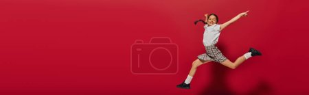 Photo for Positive and stylish preadolescent girl in white t-shirt and plaid skirt jumping and pointing with finger on red background, hairstyle and trendy accessories concept, banner - Royalty Free Image