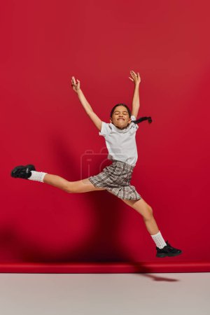 Podekscytowany i wesoły nastolatek dziewczyna w białym t-shirt i stylowe kratkę spódnica skoki i zabawy podczas pozowania na czerwonym tle, fryzura i modne akcesoria koncepcji