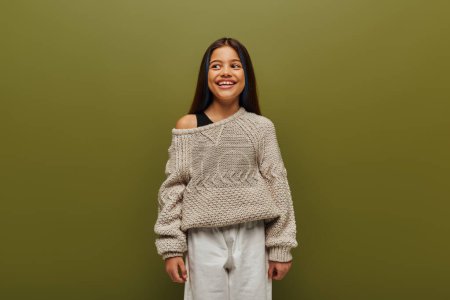 Photo for Gleeful and stylish brunette preteen girl with dyed hair wearing knitted sweater and looking away while standing and posing isolated on green, relaxed autumn vibes concept - Royalty Free Image