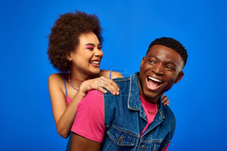 Smiling young african american woman with bold makeup hugging trendy best friend in summer outfit and spending time together isolated on blue, best friends in matching outfits, friendship
