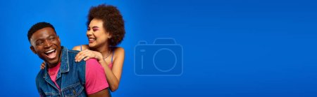 Fröhliche junge afrikanisch-amerikanische Frau mit kühnem Make-up umarmt stilvolle beste Freundin im Sommer-Outfit, während sie Spaß hat und isoliert auf blau steht, Banner, beste Freunde in passenden Outfits