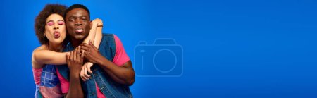 Photo for Young african american best friends in bright and trendy summer outfits hugging and sticking out tongues while standing together isolated on blue, banner, best friends in matching outfits - Royalty Free Image