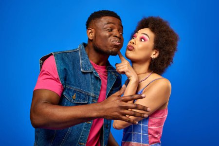 Young african american woman with bold makeup touching cheek of best friend in stylish summer outfit while standing together and having fun isolated on blue, best friends in matching outfits