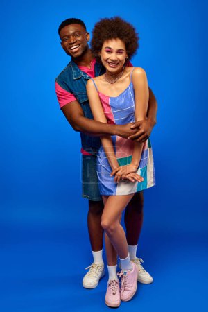 Full length of smiling african american man in bright summer outfit hugging stylish best friend in sundress while standing together on blue background, best friends in matching outfits