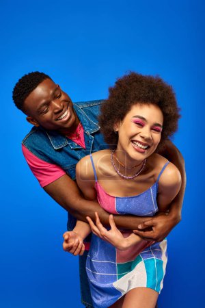 Positive afrikanisch-amerikanische Mann in hellen Sommer-Outfit umarmt stilvolle beste Freundin mit kühnem Make-up und Sundress, während sie zusammen isoliert auf blau stehen, beste Freunde in passenden Outfits