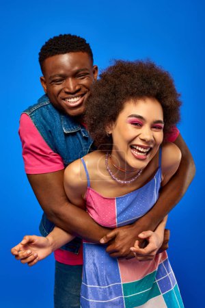 Cheerful young african american man in bright summer outfit hugging stylish best friend with bold makeup and sundress while standing isolated on blue, best friends in matching outfits
