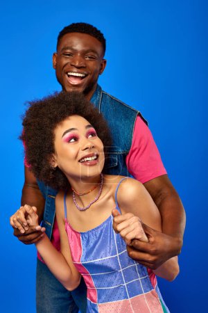Positive junge afrikanisch-amerikanische Mann in hellen Sommer-Outfit hält Hände von stilvollen besten Freund mit kühnem Make-up, während sie isoliert auf blau stehen, beste Freunde in passenden Outfits, Freundschaft