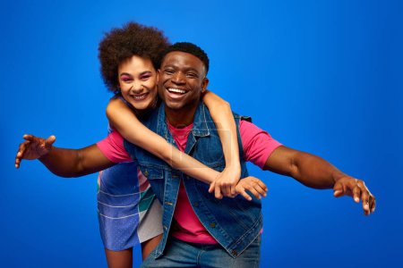 Cheerful young african american woman with bold makeup and sundress hugging fashionable best friend in summer outfit while standing together isolated on blue, best friends having good time