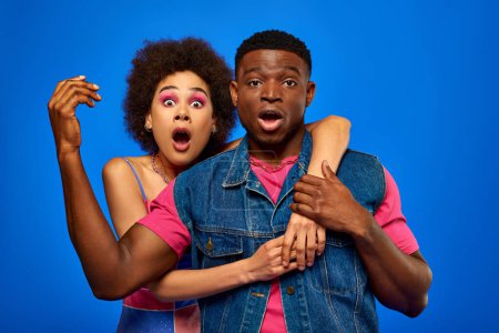 Shocked young african american woman with bold makeup hugging best friend in bright summer outfit while looking at camera and standing isolated on blue, fashionable friends in trendy clothes