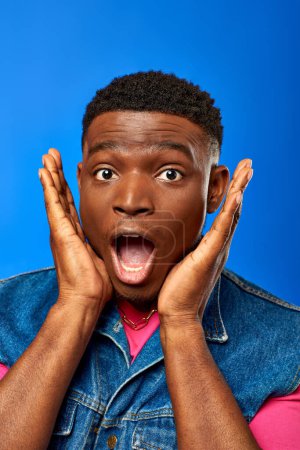 Portrait eines erstaunten jungen afrikanisch-amerikanischen Mannes mit moderner Frisur, der Jeansweste und rosa T-Shirt trägt, während er isoliert in die Kamera blickt und einen blauen, trendigen Mann zeigt, der Sommerstil zeigt
