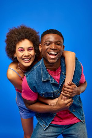 Joyeux jeune femme afro-américaine avec un maquillage audacieux en robe de soleil embrassant élégant meilleur ami en gilet en jean et en regardant la caméra isolée sur bleu, amis à la mode dans des vêtements à la mode