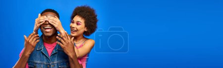 Smiling young african american woman with bright makeup covering eyes of trendy best friend in denim vest and standing isolated on blue, fashionable friends in trendy clothes, banner 