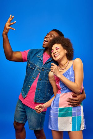Cheerful young african american man in denim vest and pink t-shirt hugging best friend in sundress and gesturing while standing isolated on blue, fashionable friends in trendy clothes