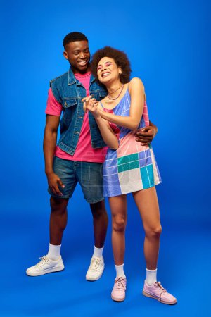 Full length of cheerful and stylish young african american man in denim vest and bright t-shirt hugging best friend in sundress on blue background, fashionable friends in trendy clothes