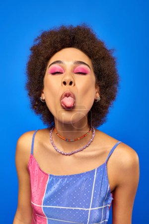 Retrato de mujer afroamericana joven y elegante con maquillaje audaz que lleva vestido de verano y sobresale de la lengua mientras está de pie aislado en azul, modelo carismático en traje de verano
