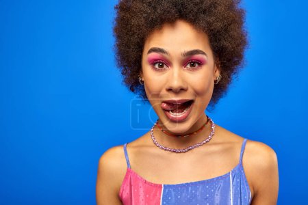 Retrato de una mujer afroamericana joven y elegante con cabello natural y maquillaje audaz que sobresale de la lengua y mira a la cámara mientras está de pie aislado en azul, modelo carismático en traje de verano