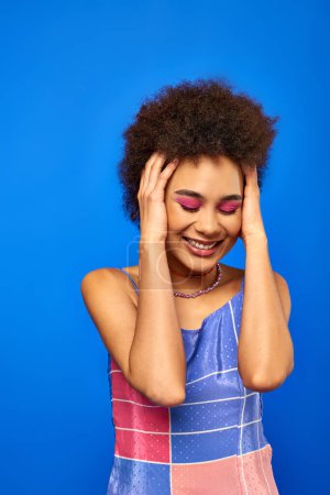 Portrait of smiling stylish young african american woman with natural hair and bold makeup touching head while standing and posing isolated on blue, charismatic model in summer outfit