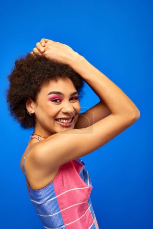 Porträt einer zufriedenen jungen afrikanisch-amerikanischen Frau mit kühnem Make-up in Sommerkleidung, während sie isoliert auf blauem, charismatischem Modell im Sommeroutfit posiert und in die Kamera blickt