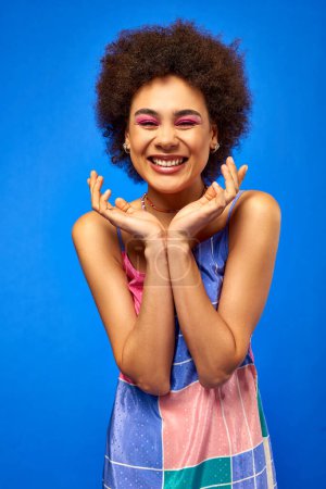 Retrato de mujer afroamericana joven y positiva con cabello natural y maquillaje audaz usando vestido de verano y posando mientras está de pie aislado en azul, modelo carismático en traje de verano