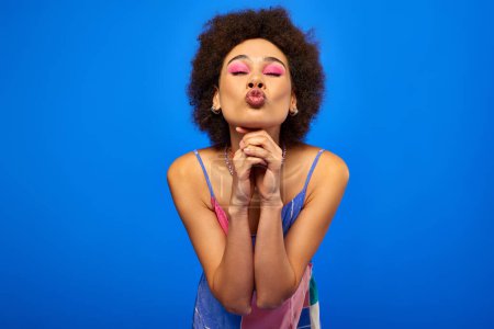 Portrait de jeune femme afro-américaine avec un maquillage élégant et audacieux portant une robe de soleil et des lèvres boudantes tout en posant et debout isolé sur un modèle bleu charismatique en tenue d'été