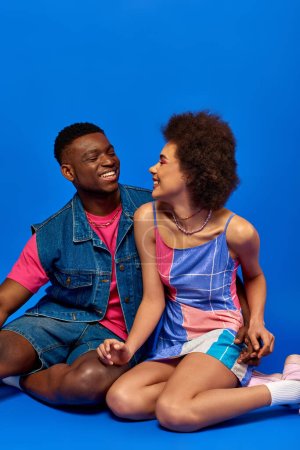 Young and stylish african american best friends in summer outfits smiling while looking at each other, spending time together and sitting on blue background, stylish friends posing confidently