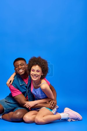 Smiling young african american man in denim vest hugging best friend with bold makeup and stylish sundress and sitting together on blue background, stylish friends posing confidently