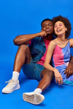 Young and stylish african american man in summer outfit grimacing while sitting near cheerful best friend in summer dress while posing on blue background, stylish friends posing confidently