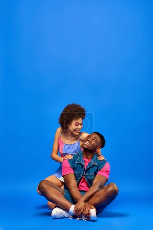Smiling young african american woman with bright makeup wearing summer dress and hugging best friend in denim vest while sitting on blue background, stylish friends posing confidently