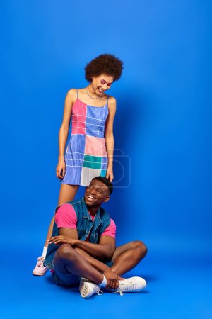 Full length of cheerful young african american man in summer outfit sitting near best friend in sundress standing on blue background, stylish friends posing confidently, friendship concept