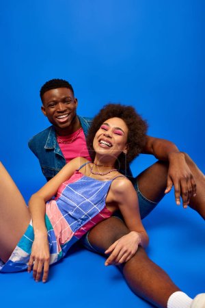Fröhliche und trendige junge afrikanisch-amerikanische beste Freunde in hellen Sommeroutfits, die vor blauem Hintergrund gemeinsam in die Kamera schauen, modische Bestien, die Zuversicht ausstrahlen 